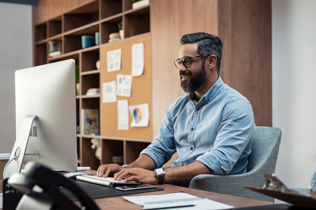 Smiling creative business man typing on desktop computer in office. Mature middle eastern businessman working at modern office space. Happy hispanic man wearing casual and eyeglasses working on his computer.