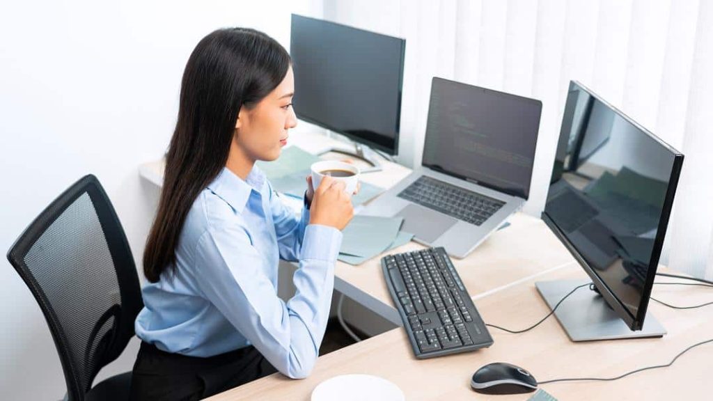 A female professional drinks a cup of coffee while looking at data from three monitors